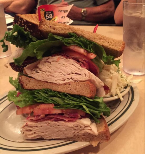 A towering sandwich with layers of turkey, lettuce, tomato, and bacon on whole grain bread, served with a side of coleslaw.