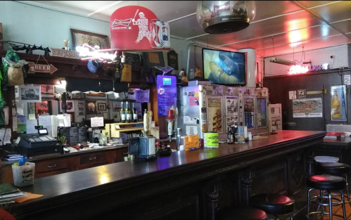 A cozy bar interior featuring a wooden counter, stools, neon signs, and various bottles and memorabilia on display.