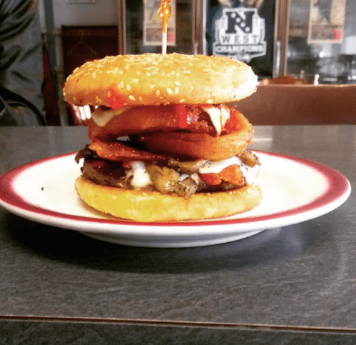 A towering burger with a sesame seed bun, layers of meat, onion rings, and sauces on a plate.