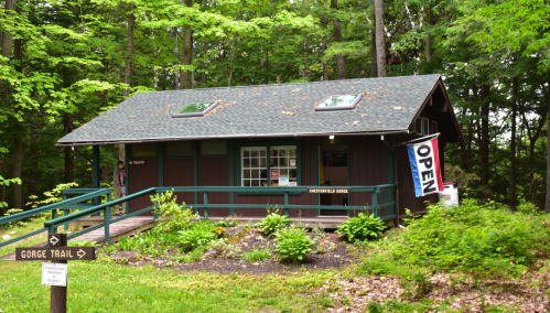 A small brown cabin in a forest, with a sign reading "OPEN" and a trail sign pointing to the Gorge Trail.