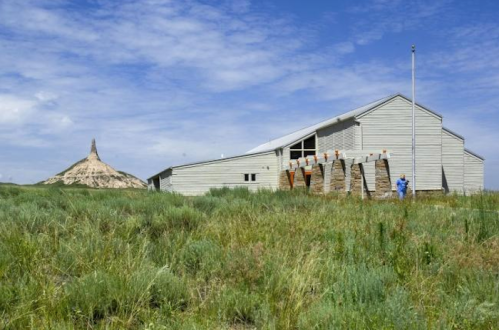 A wooden building beside a grassy field, with a distinctive rock formation in the background under a blue sky.