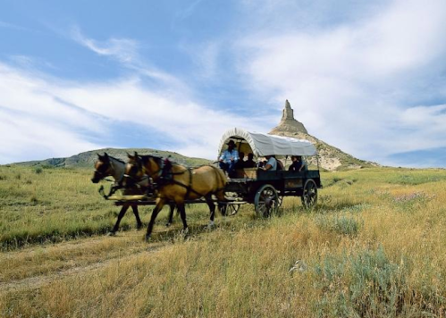 A horse-drawn wagon travels through a grassy landscape with a rocky formation under a blue sky.