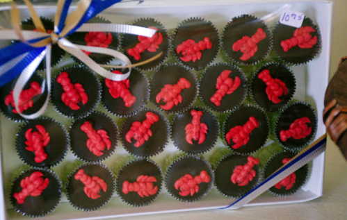 A box of chocolate cupcakes topped with red lobster-shaped candies, decorated with blue and white ribbons.