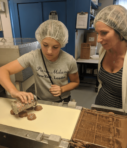 A girl in a hairnet sprinkles colorful toppings on chocolate treats while an adult watches attentively.