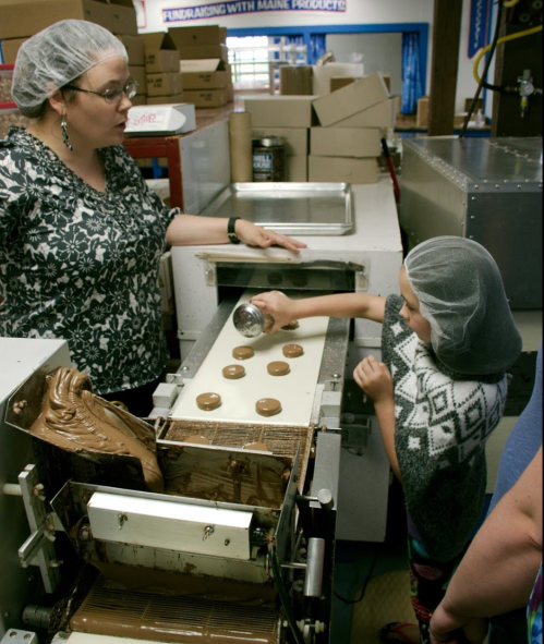 A woman and a child in hairnets pour chocolate onto a conveyor belt in a candy production facility.