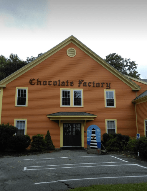 Orange building with "Chocolate Factory" sign, featuring a front door and landscaped area with trees and shrubs.