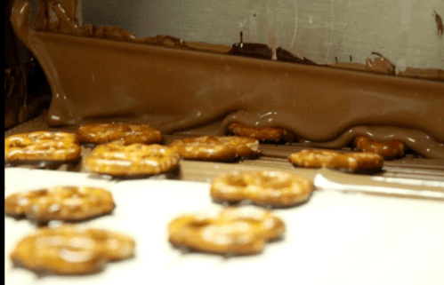 Pretzels being coated in flowing chocolate on a conveyor belt in a factory setting.