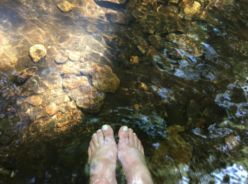 Bare feet in clear water, surrounded by pebbles and sunlight reflecting on the surface.