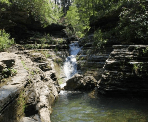 A serene waterfall cascades over rocky cliffs, surrounded by lush greenery and a clear pool of water below.