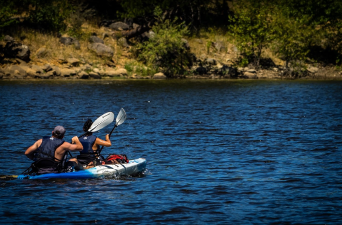 Two people kayaking on a calm river, paddling away from the camera with trees and rocks in the background.