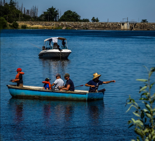 A group of people fishing from a small boat on a calm lake, with another boat in the background.