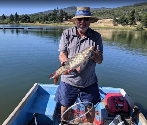 A man in a straw hat holds a large fish while standing in a small boat on a calm lake surrounded by trees.