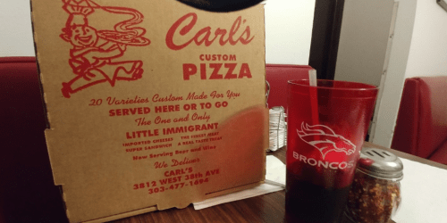 A pizza box from Carl's Custom Pizza next to a red cup and a shaker on a table in a restaurant.