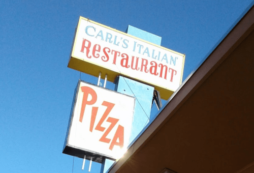Sign for Carl's Italian Restaurant featuring "Pizza" in bold red letters against a clear blue sky.