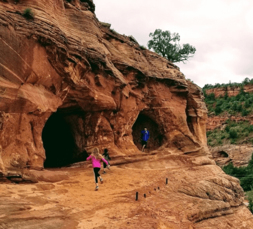 Two people explore rocky caves in a red sandstone landscape, surrounded by greenery and cloudy skies.