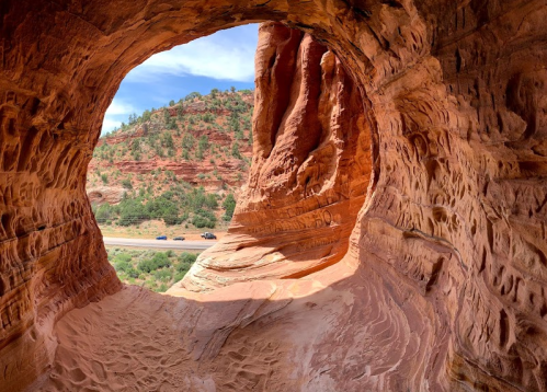 View through a natural rock arch, showcasing red sandstone formations and a distant road surrounded by greenery.
