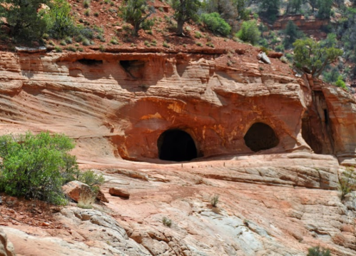 Two dark cave openings in a red rock formation, surrounded by greenery and rugged terrain.