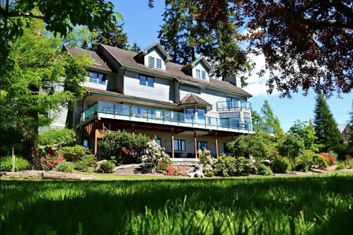 A large, multi-story house surrounded by lush greenery and colorful flowers under a clear blue sky.