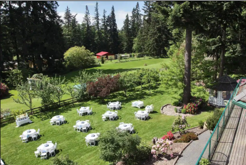 A scenic outdoor venue with white tables set on green grass, surrounded by trees and colorful flowers.