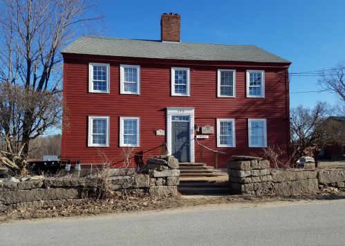 A red two-story house with white windows and a stone wall in front, set against a clear blue sky.