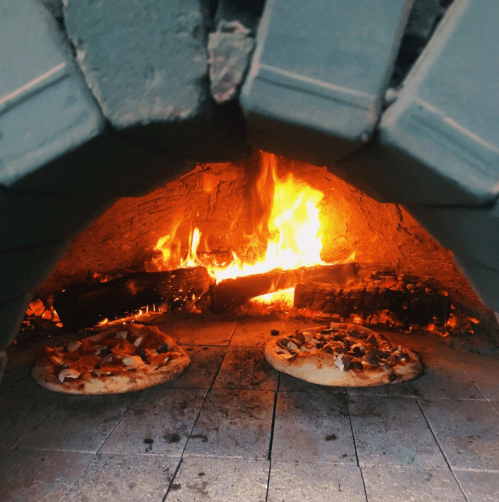 Two pizzas baking in a wood-fired oven, with flames and glowing embers visible in the background.