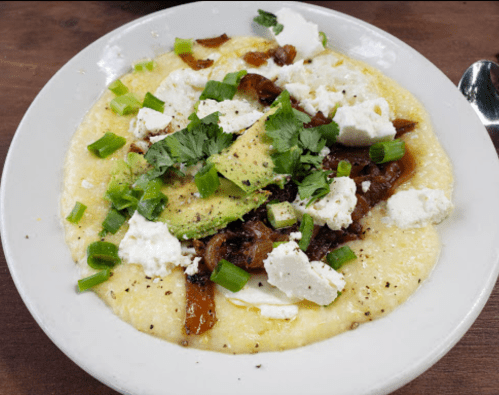 A bowl of creamy grits topped with avocado, pulled meat, cheese, green onions, and cilantro.