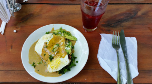 A plate of eggs topped with sauce and greens, accompanied by a drink and utensils on a wooden table.