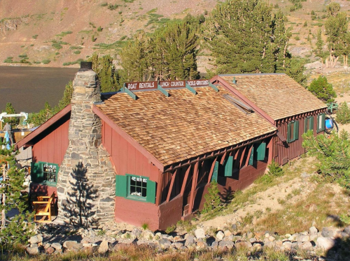 A rustic wooden building with a stone chimney, surrounded by trees and mountains, near a lake. Boat rentals sign visible.