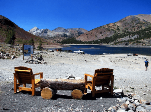 Two wooden chairs facing a serene lake surrounded by mountains, with a clear blue sky above.