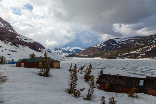 A serene snowy landscape with mountains, a frozen lake, and rustic cabins under a cloudy sky.