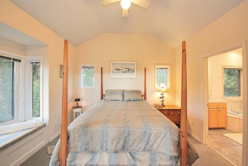 Cozy bedroom with a four-poster bed, soft blue bedding, and natural light from large windows. Bathroom visible in the background.