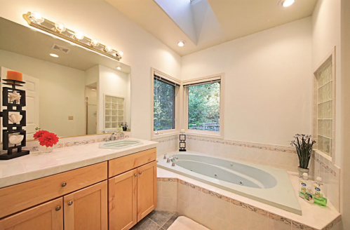 Bright bathroom featuring a soaking tub, wooden cabinetry, and large windows with a view of greenery outside.