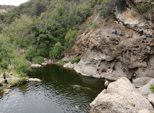 A scenic outdoor area with a swimming hole surrounded by rocky cliffs and people enjoying the water and nature.