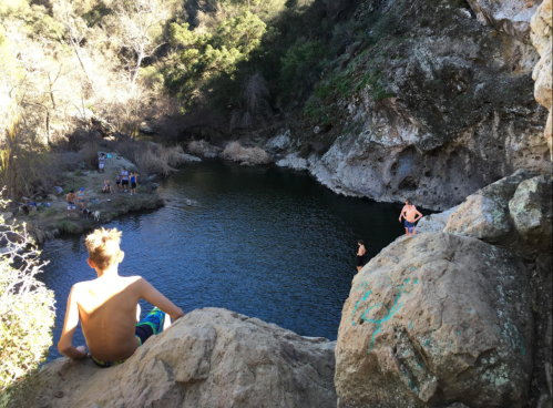 A boy sits on a rock overlooking a serene swimming hole surrounded by trees and people enjoying the water.