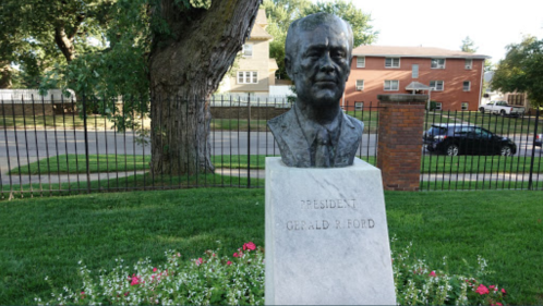A bust of President Gerald R. Ford on a stone pedestal, surrounded by grass and flowers, with a fence and buildings in the background.