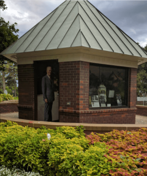 A small brick building with a glass window, surrounded by colorful plants, featuring a life-sized cutout of a man inside.