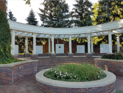 A circular memorial structure with columns, surrounded by greenery and plaques, set in a peaceful outdoor area.