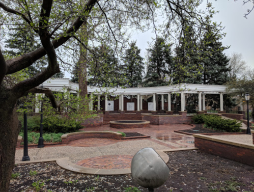 A landscaped park featuring a white gazebo, brick pathways, and greenery, surrounded by tall trees on a cloudy day.
