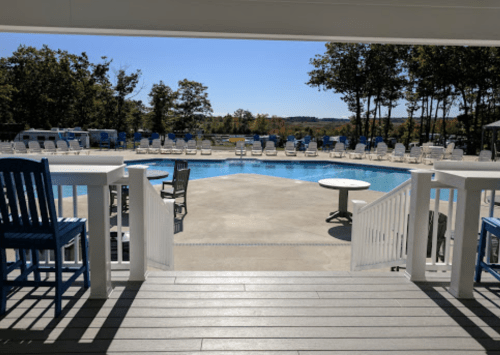 View of a sunny pool area with blue chairs and tables, surrounded by trees and a clear sky.
