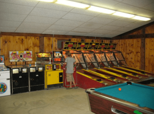 A retro arcade with pinball machines, a pool table, and vintage game cabinets in a wooden-paneled room.