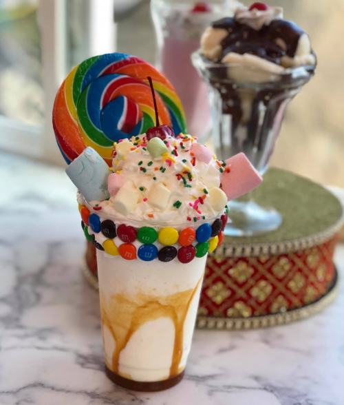 A colorful milkshake topped with whipped cream, a rainbow lollipop, marshmallows, and candy.