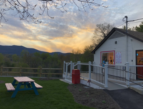 A white building with a red door overlooks a scenic landscape at sunset, featuring a picnic table in the foreground.