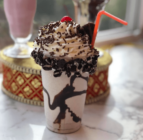 A decadent milkshake topped with whipped cream, chocolate syrup, sprinkles, and an Oreo cookie, served in a clear cup.