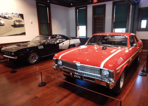 Two classic cars on display: a black muscle car and a red 1970 Chevrolet Nova, set in a well-lit museum interior.