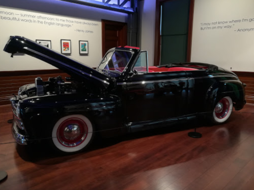 A vintage black convertible car with the hood open, displayed in a gallery setting.