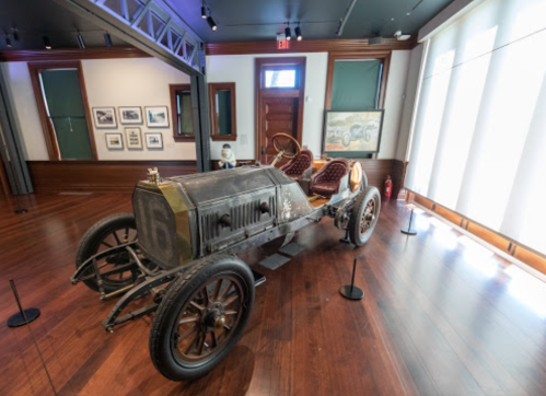 A vintage car displayed in a museum, featuring wooden floors and framed photographs in the background.