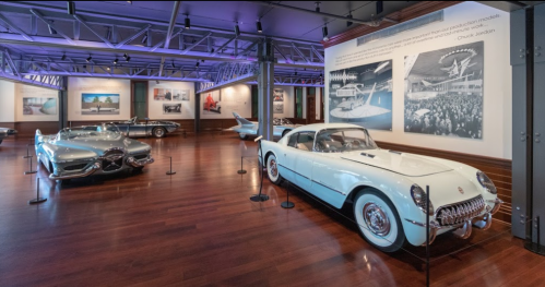A museum exhibit featuring classic cars, including a light blue Corvette and a futuristic silver vehicle, with photos on the wall.