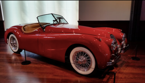 A classic red convertible car with whitewall tires, displayed indoors on a wooden floor.