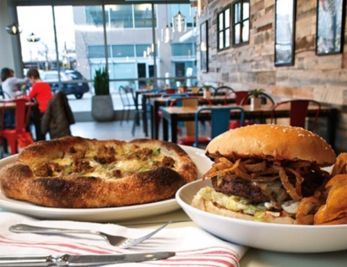 A burger with crispy onions and a side of sweet potato fries, alongside a cheesy flatbread, in a cozy restaurant setting.