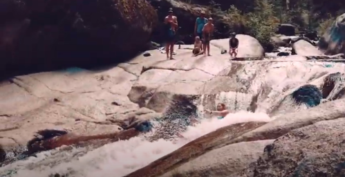 A group of people enjoying a natural water slide on smooth rocks, surrounded by trees and boulders.